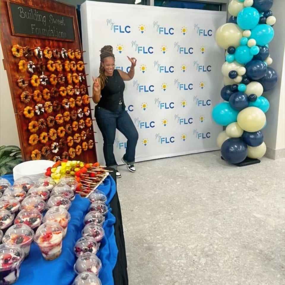 Woman poses beside a dessert table, flower wall, branded backdrop, and blue, teal, white, and navy balloon display.