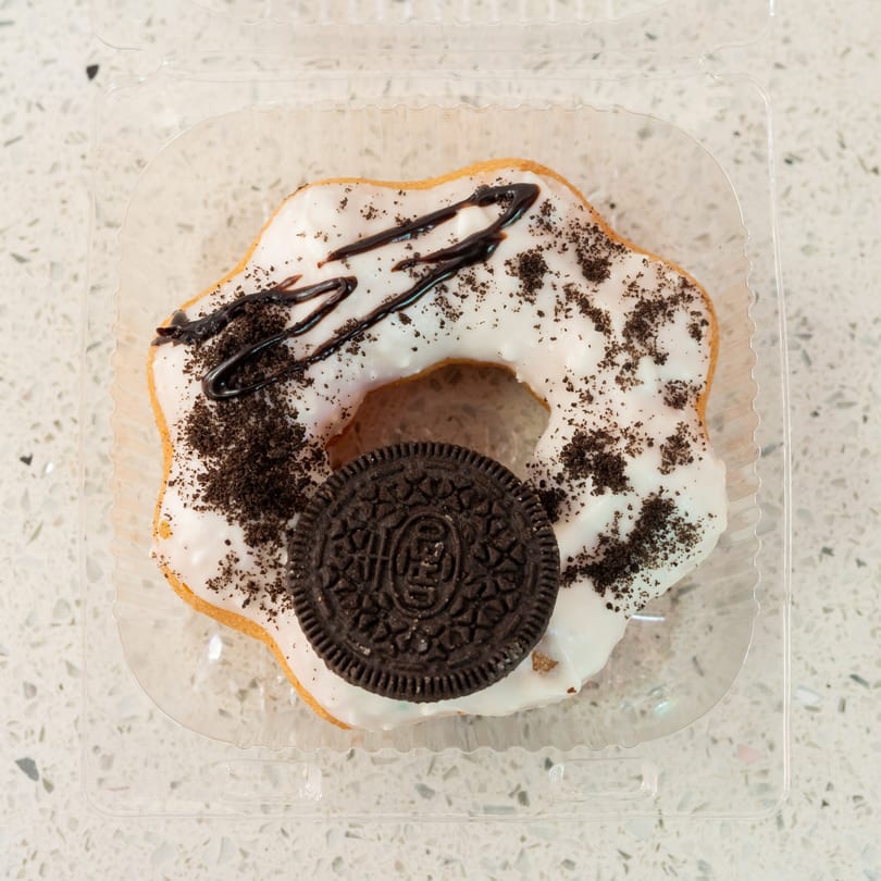 White-frosted donut with chocolate drizzle, cookie crumbles, and a whole cookie in a plastic container.