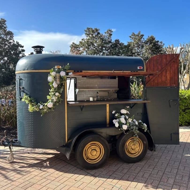 Dark green food truck with gold trim, decorated with white flowers, parked on a brick patio.
