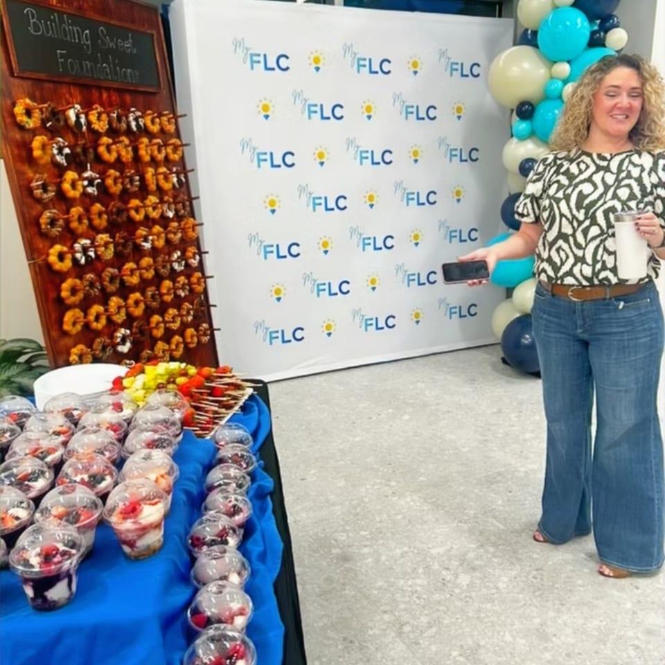 Woman holds a white cup beside a dessert table, balloon decorations, and a backdrop reading “My FLC.”