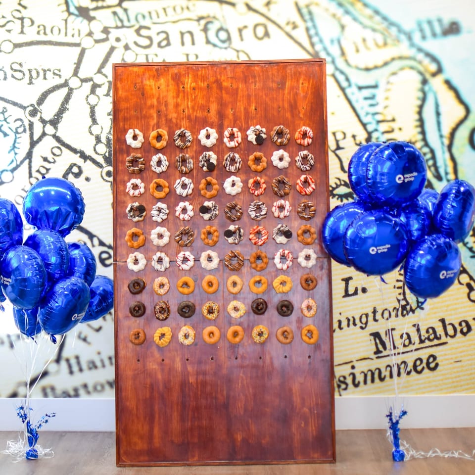 Wooden display board showcases rows of decorated doughnuts, surrounded by blue balloons against a San Francisco map backdrop.