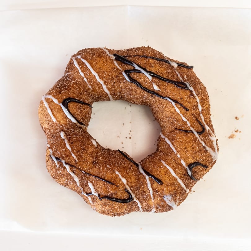 A large, irregularly shaped churro donut covered in cinnamon sugar, drizzled with white and dark icing.