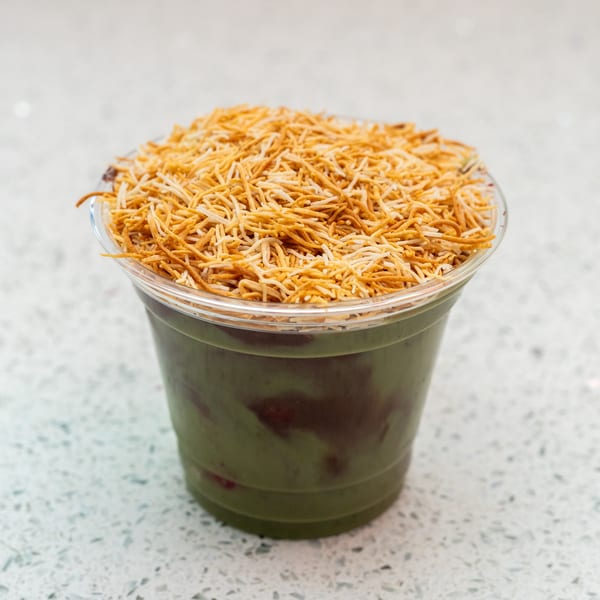 Green smoothie with red swirls, topped with crispy golden noodles in a clear plastic cup.