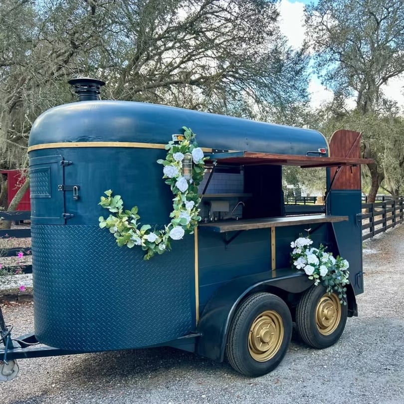 A dark blue food trailer with gold trim and wheels, decorated with white flowers, parked on a gravel path.