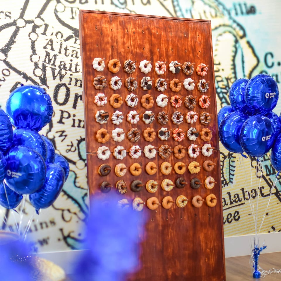 Decorated doughnuts arranged on a wooden pegboard, surrounded by blue balloons against a colorful illustrated backdrop.