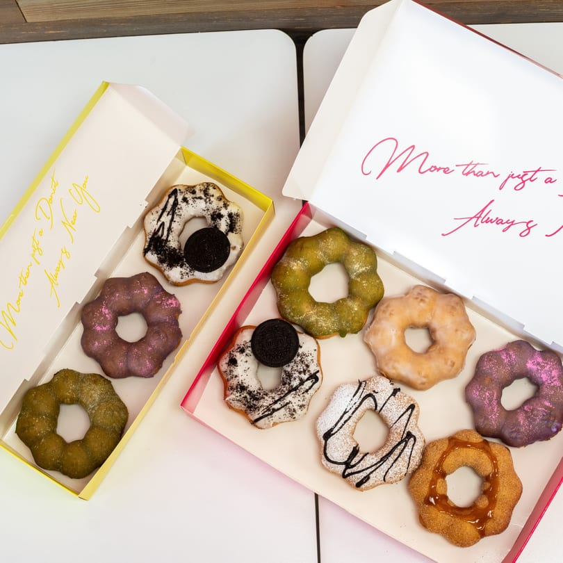 Two open boxes of colorful, uniquely shaped donuts with various toppings on a white table.