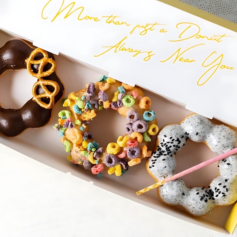 Three colorful mochi donuts, including chocolate pretzel, cereal, and black sesame, in a white box.
