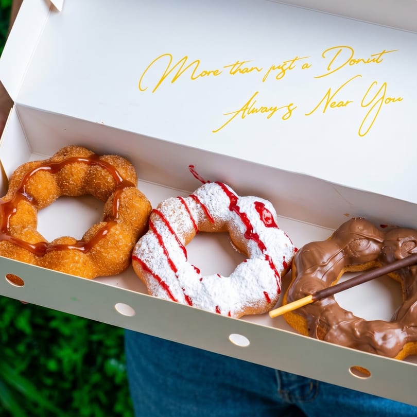 A white box holds three mochi donuts: caramel, powdered sugar with raspberry, and chocolate with a Pocky stick.