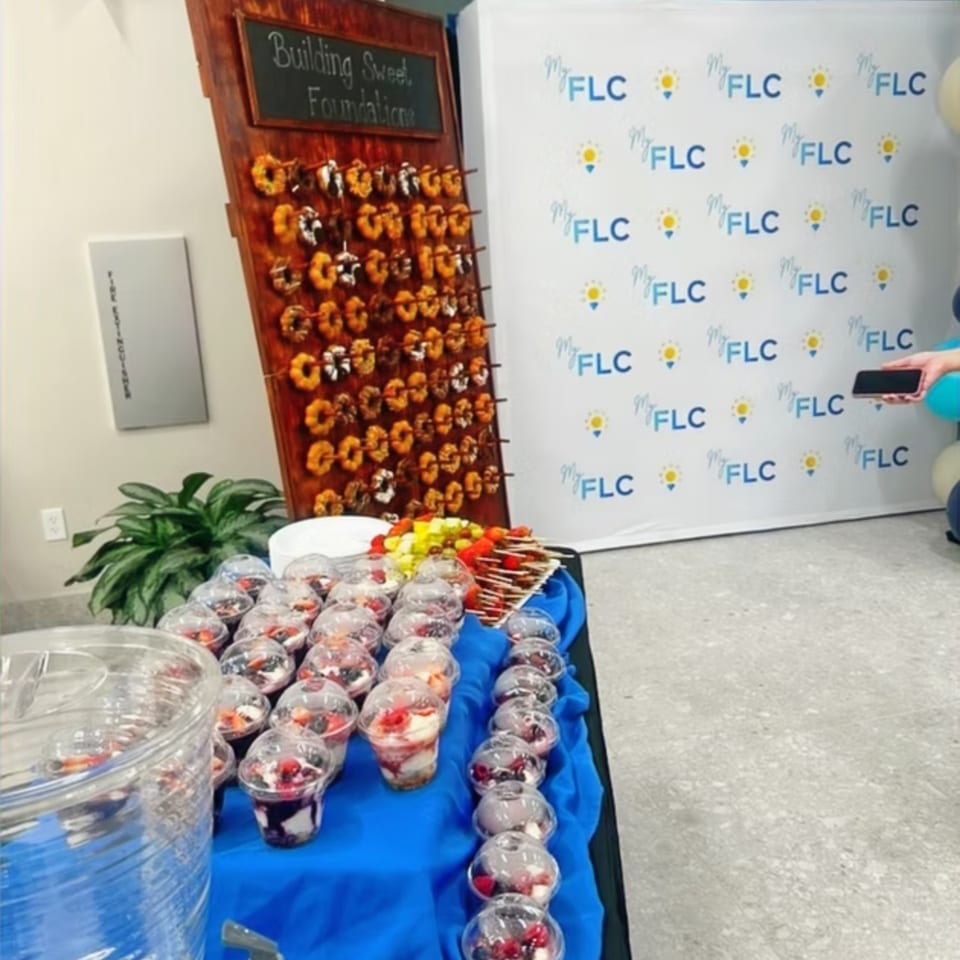Catering table displays individual fruit parfait cups, dessert skewers, and a colorful snack display against an FLC-branded backdrop.