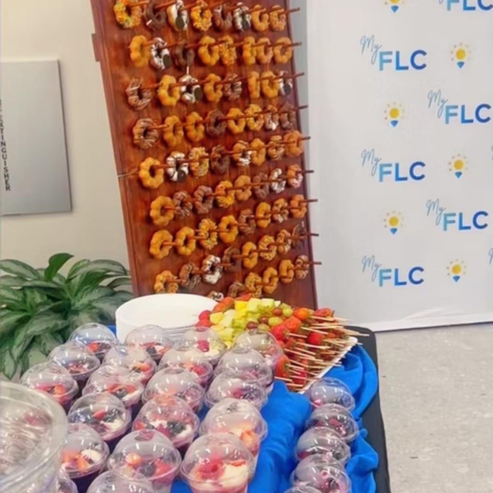 A buffet display features rows of grilled shrimp skewers, colorful fruit cups, and salad in front of a branded backdrop.